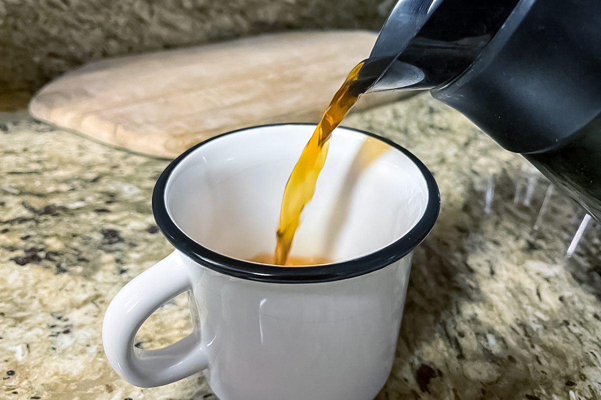 A stream of coffee is being poured from a black pot into a white enamel mug with a black rim, sitting on a granite countertop. A wooden cutting board is in the background.
