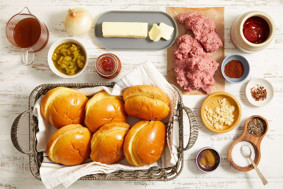 A top view of ingredients for making sloppy joes, including six sandwich buns, ground beef, butter, onion, ketchup, mustard, pickles, spices, sugar, and broth, all arranged neatly on a white surface.