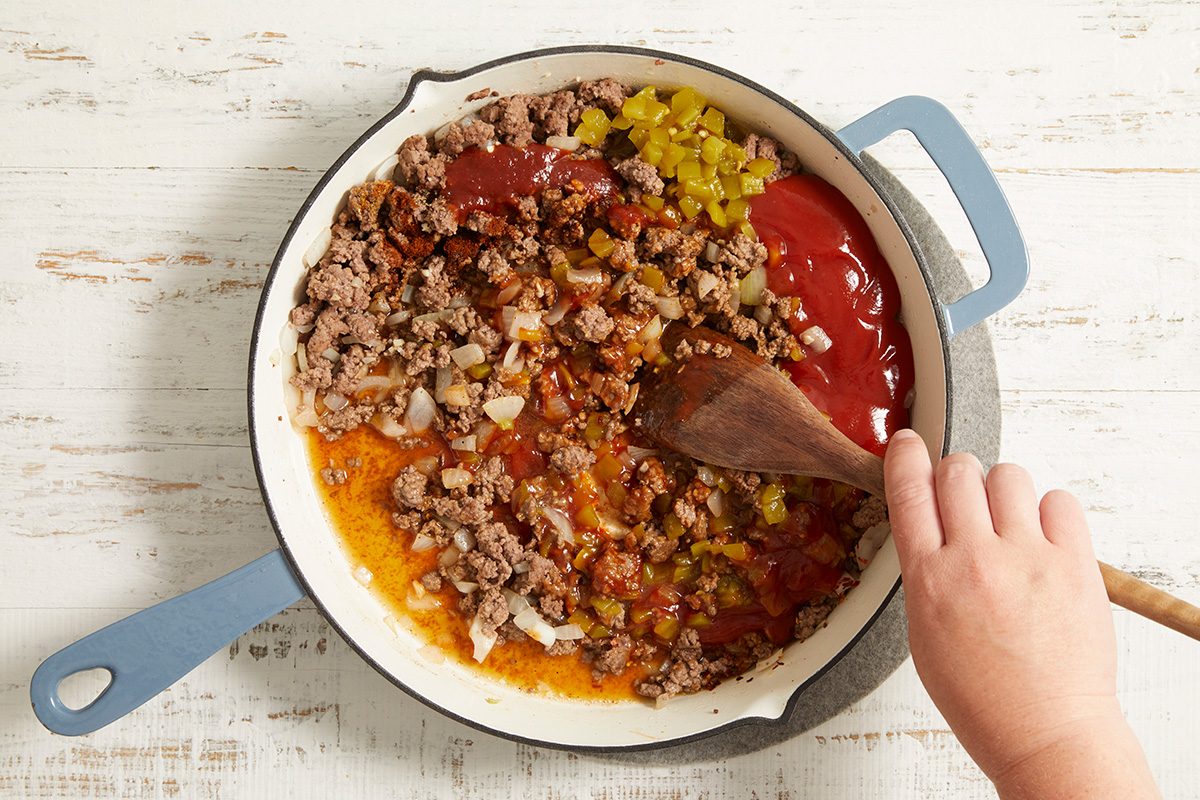 A hand stirs ground beef, chopped onions, ketchup, relish, and seasonings in a white skillet with a blue handle on a white wooden surface.