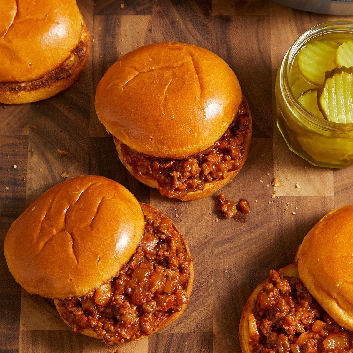 Four sloppy joe sandwiches with soft buns are arranged on a wooden surface, next to an open jar of pickle slices.