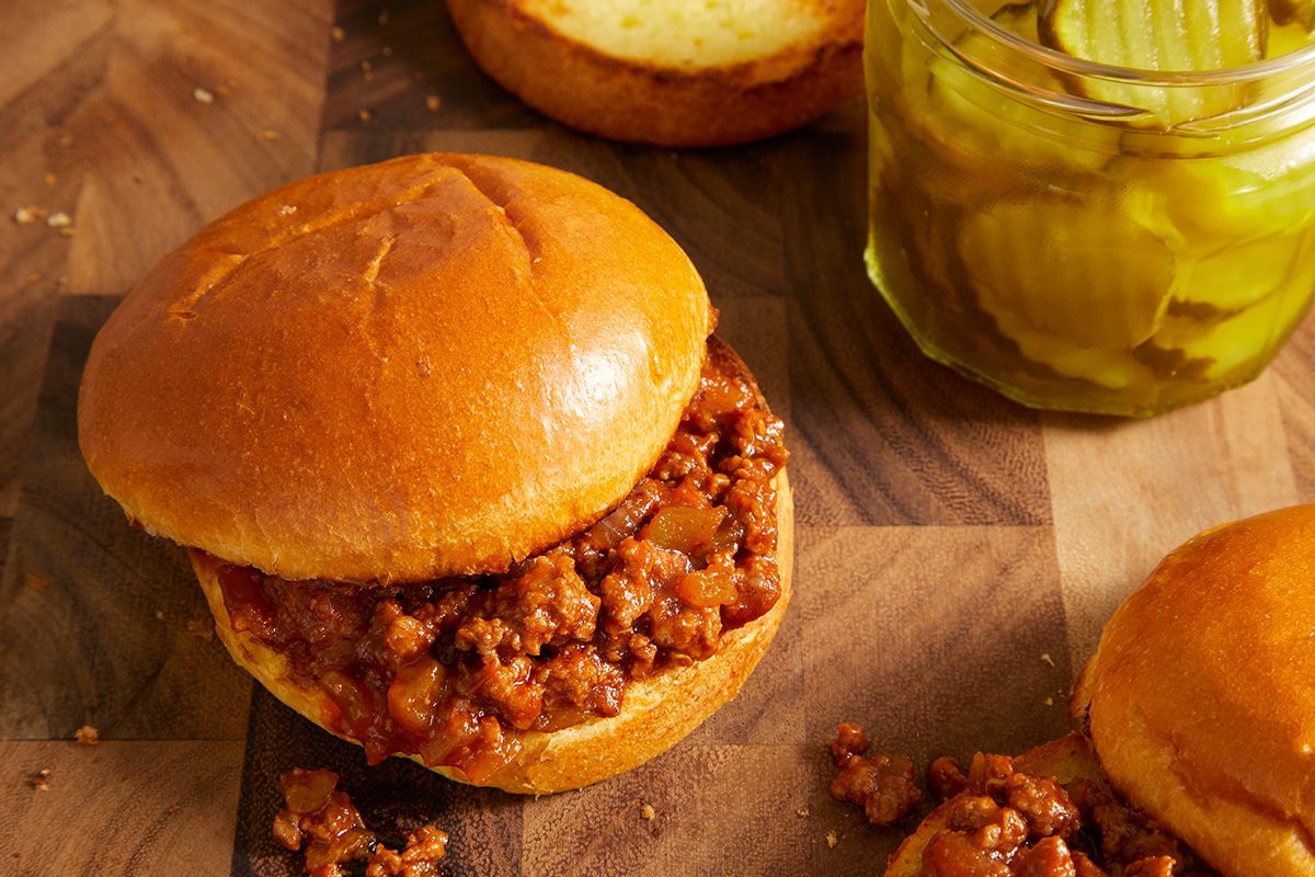 A sloppy joe sandwich on a wooden surface next to a glass jar of pickle slices, with a sandwich bun top and some filling spilled nearby.