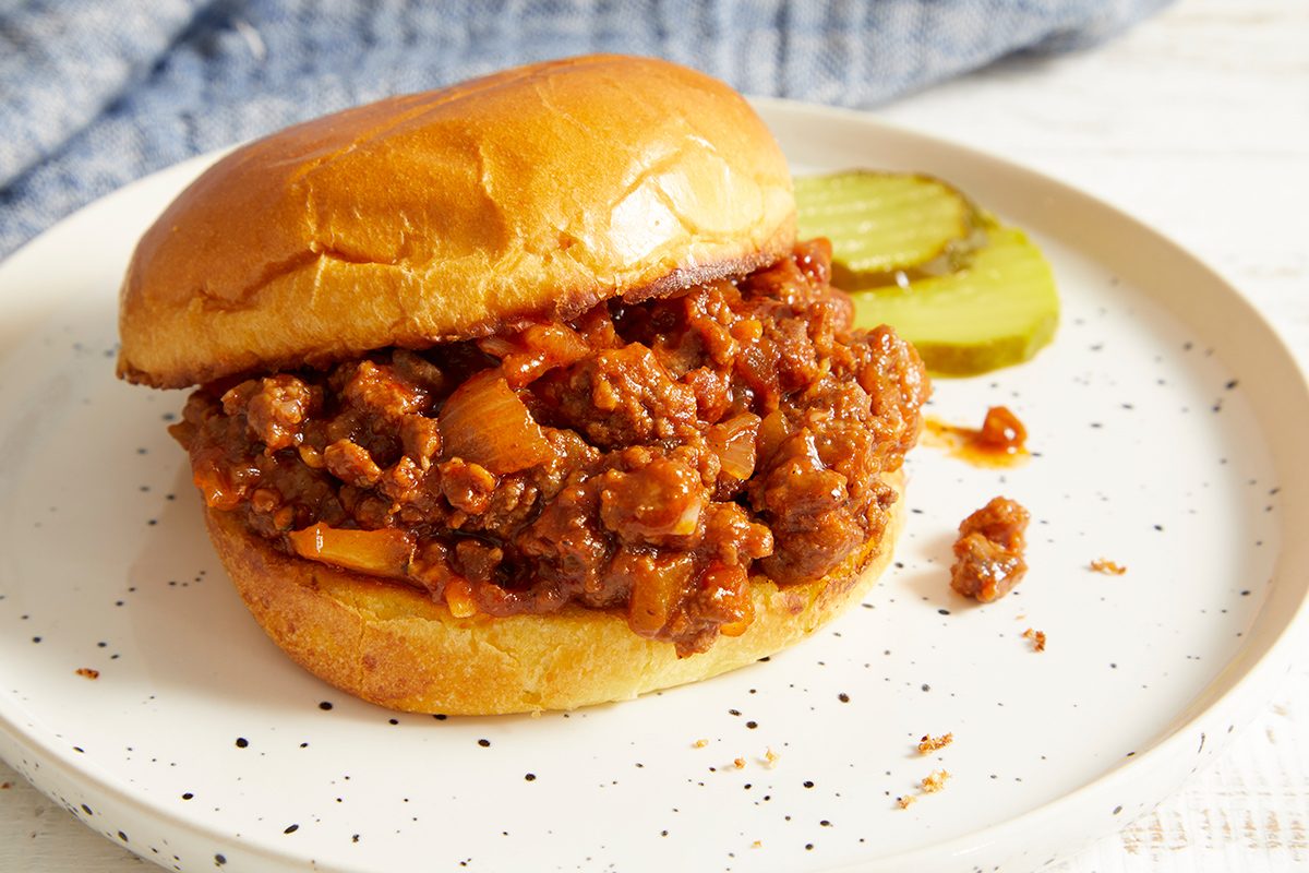 A sloppy joe sandwich with a soft bun and savory meat filling sits on a white speckled plate, accompanied by two pickle slices on the side.