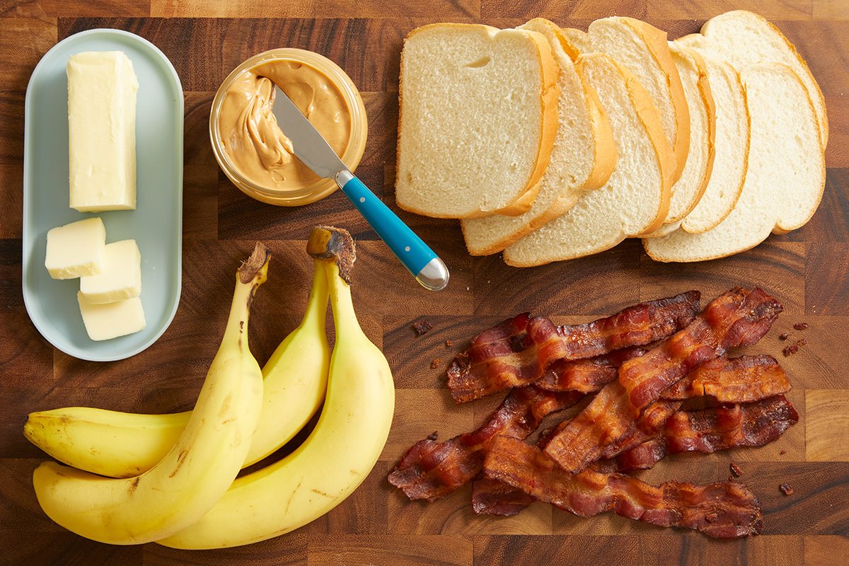 A wooden surface with sliced white bread, cooked bacon strips, two bananas, a dish of butter, and an open jar of peanut butter with a knife.