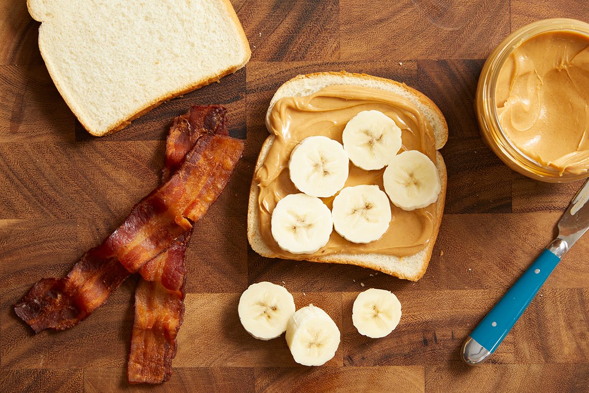 A slice of bread with peanut butter and banana slices, next to two strips of cooked bacon, a plain bread slice, a jar of peanut butter, and a knife on a wooden surface.