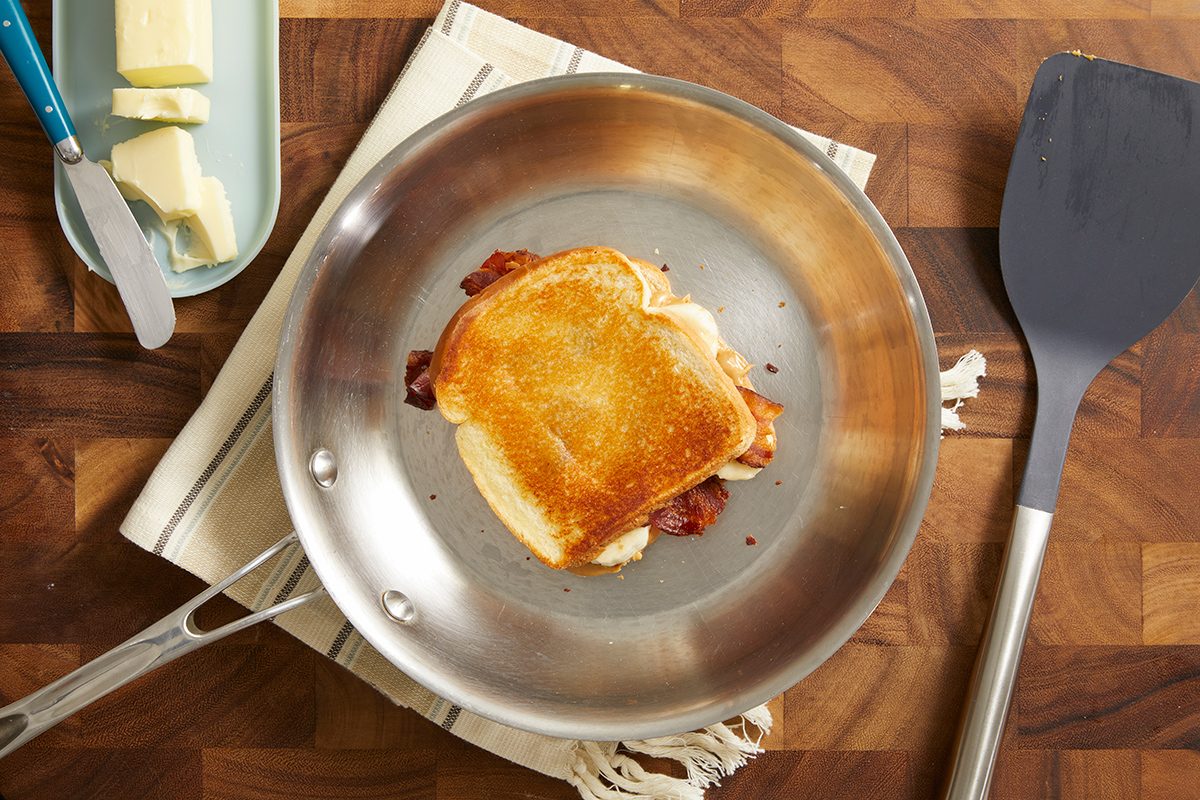 A grilled cheese sandwich cooking in a stainless steel skillet on a wooden surface, with a spatula nearby and a butter dish with a knife on the side.