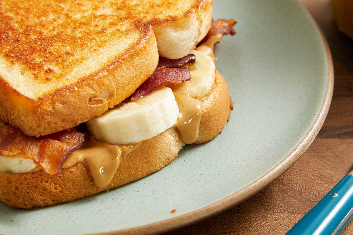 A close-up of a sandwich on a light blue plate, filled with peanut butter, banana slices, and crispy bacon between two thick slices of toasted bread.