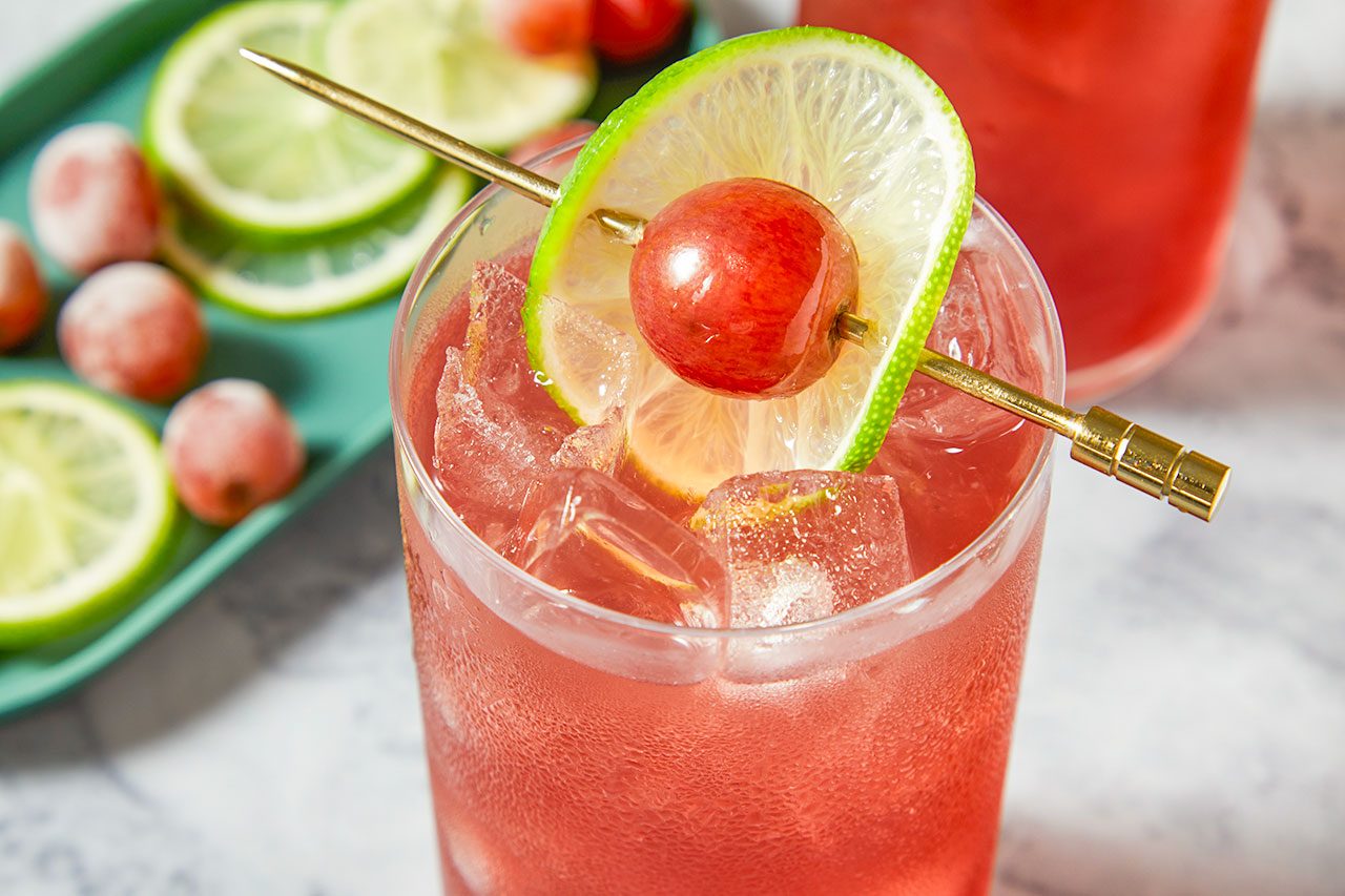 Close-up shot of a Transfusion cocktail served over ice in a highball glass, garnished with a lime wheel and a skewered red grape, highlighting the sparkling pink drink on a marble surface with fresh citrus in the background;
