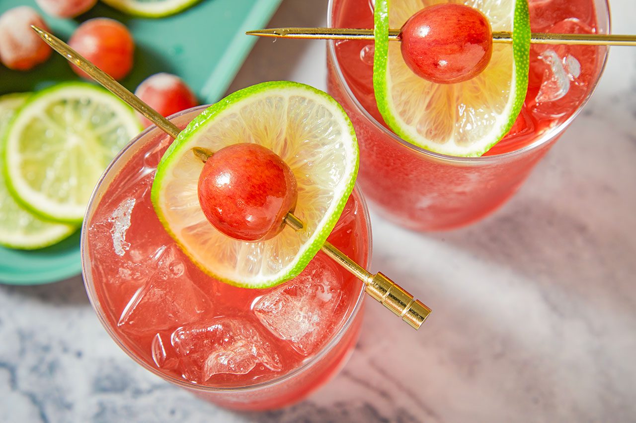 Overhead shot of two Transfusion cocktails served over ice in highball glasses, each garnished with a lime wheel and a skewered red grape, arranged on a marble surface with fresh lime slices and grapes in the background;