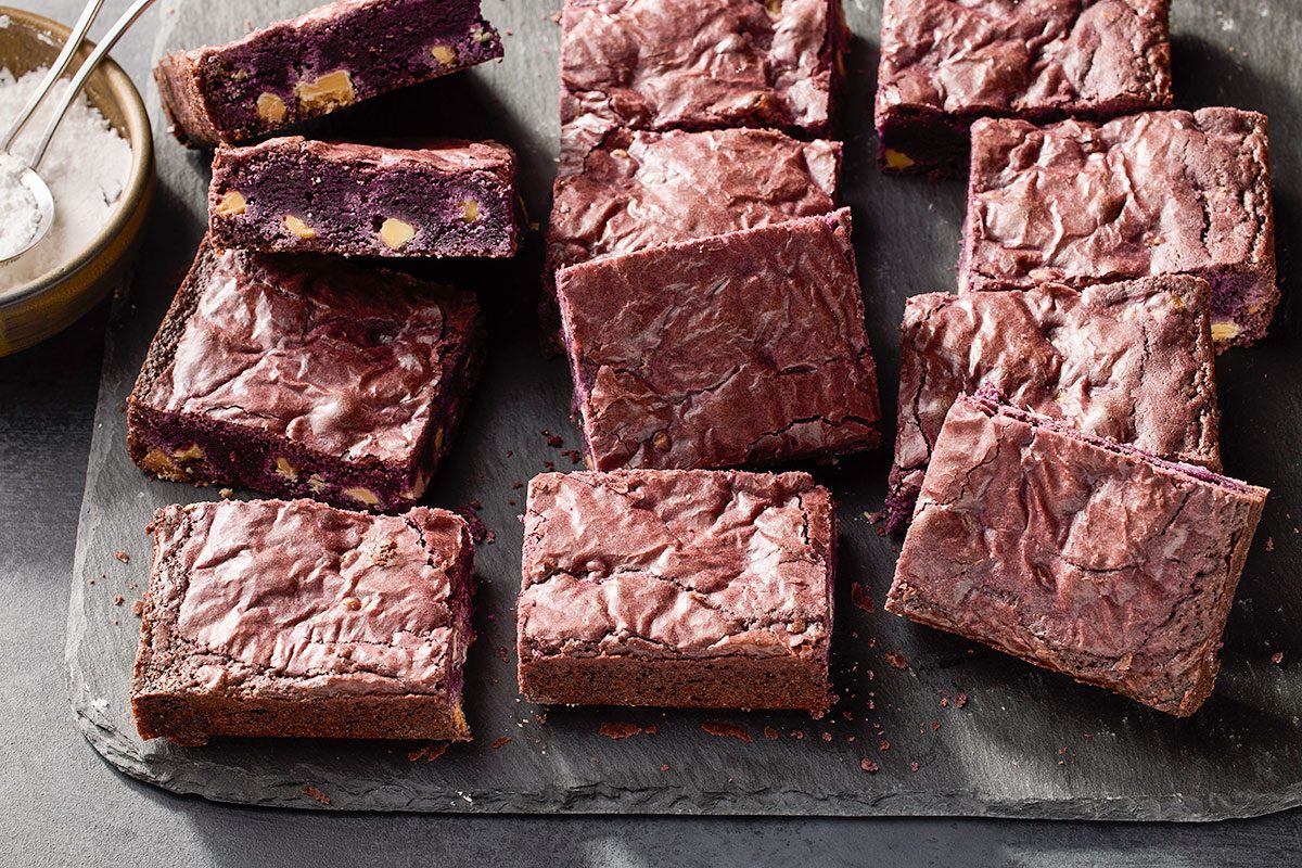 Overhead horizontal image of several square-cut purple brownies arranged on a dark slate surface. The brownies have a rich purple interior with visible white chocolate chunks, and a small bowl of powdered sugar with measuring spoons is visible in the upper left corner.