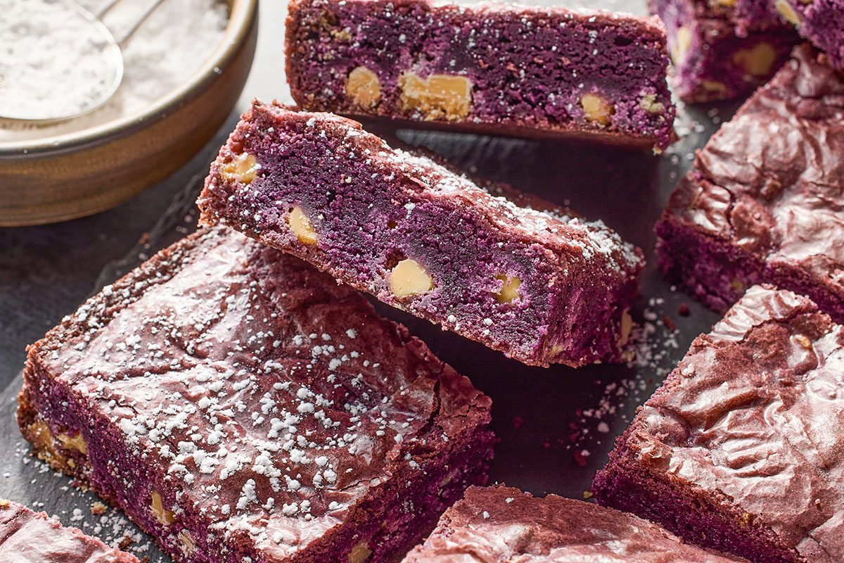 Eye Level horizontal image of several purple brownies with white chocolate chips, dusted with powdered sugar. Some brownies are stacked while others are arranged on a dark surface. In the background, a bowl of powdered sugar and a sifter are visible.