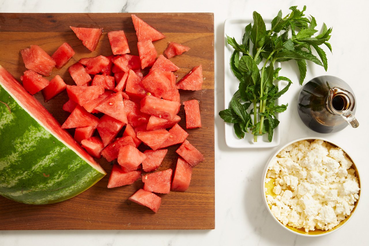 Overhead shot of cubed watermelon and a watermelon wedge arranged on a wooden cutting board, with fresh mint, crumbled feta cheese, and balsamic vinegar in small bowls and dishes nearby