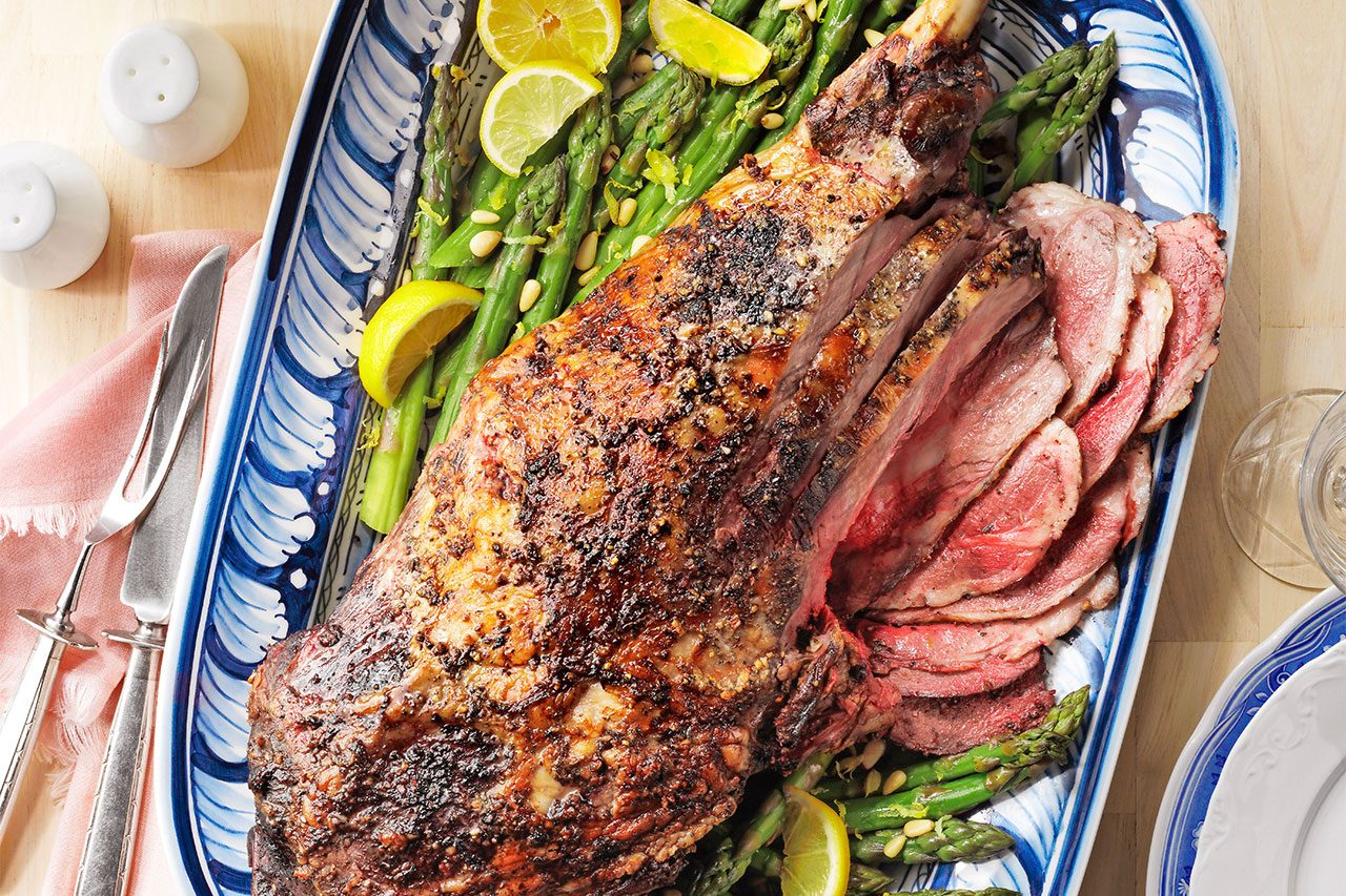 Roast Leg of Lamb; Holiday & Celebrations Cookbook 2021; Easter Dinner; overhead camera angle; pale birch wood surface; blue white platter; pink roses; white hydrangeas; white blue flower charger plate; white filigree detail dinner plate; white salt pepper shakers; light pink fringe cloth napkin; lemons; asparagus; leg of lamb; garlic cloves; dried thyme; dried marjoram; dried oregano; canola oil; roasting pan