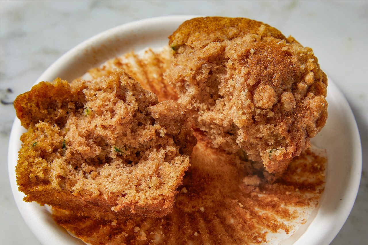 3/4 angle view shot of a zucchini coffee cake muffin split open on a small plate, revealing its moist crumb, served alongside a cup of black coffee with additional streusel-topped muffins in the background;