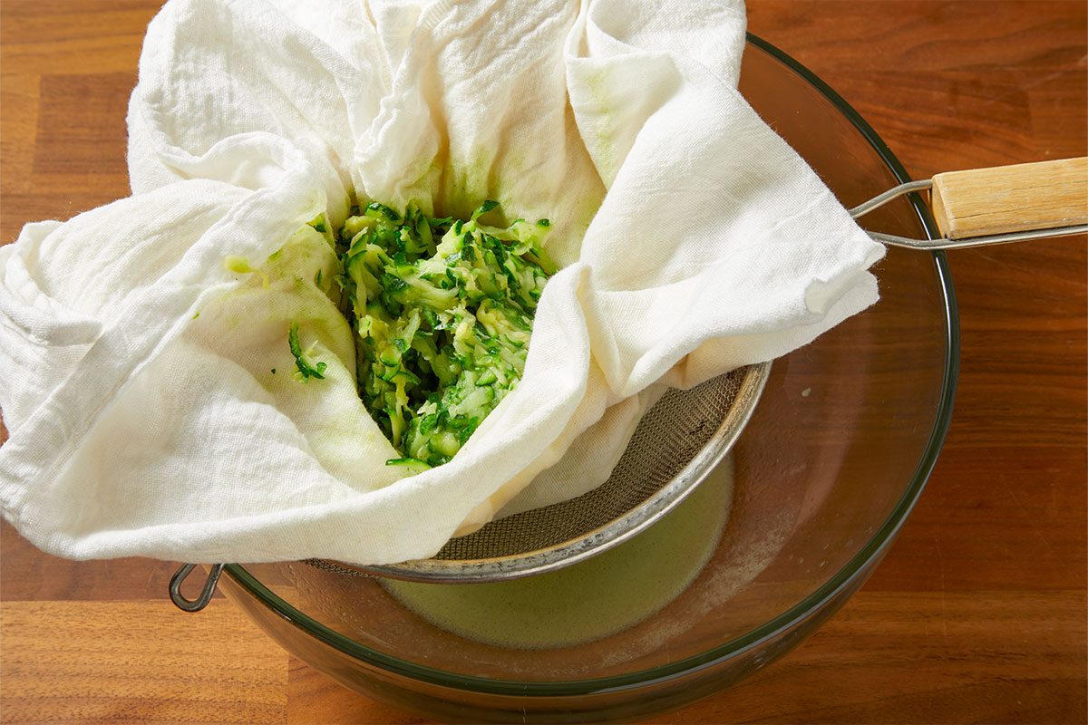 Overhead horizontal image of grated zucchini in a white cheesecloth, placed inside a metal sieve. The sieve is resting over a clear glass bowl, collecting liquid that has been squeezed from the zucchini.