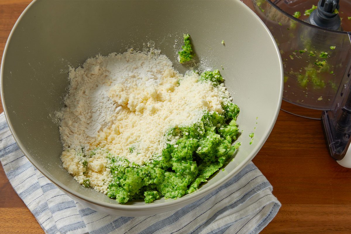 Overhead horizontal image of a gray bowl on a wooden surface, holding distinct piles of finely chopped green broccoli, white flour, and grated Parmesan cheese. A food processor containing green remnants is visible in the background, resting on a striped kitchen towel.