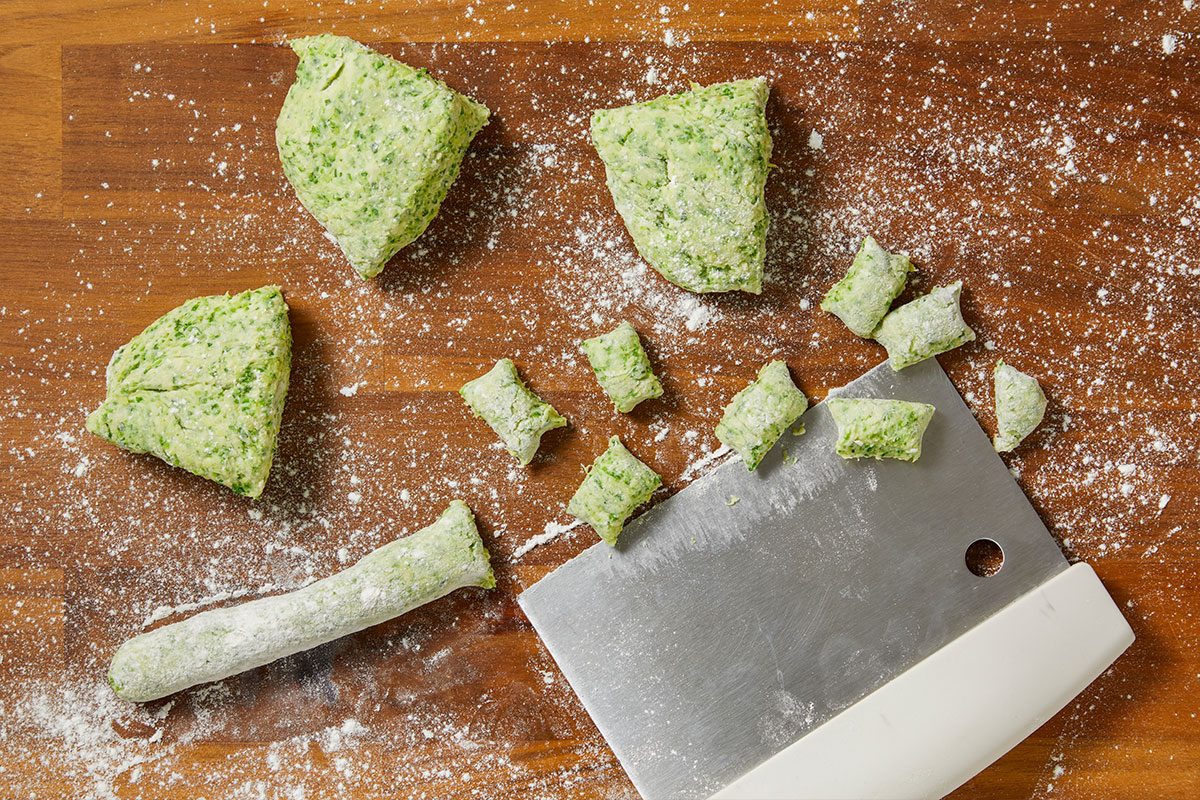 Overhead horizontal image of green dough being prepared on a wooden surface dusted with flour. Several pieces of dough are shown, including large mounds, a rolled log, and smaller gnocchi-like portions that have been cut, with a metal bench scraper nearby.