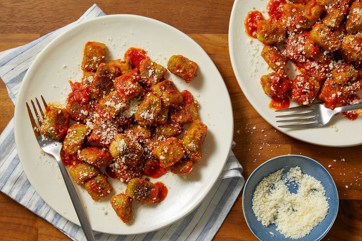 Overhead horizontal image of two white plates of green gnocchi covered in red tomato sauce and sprinkled with grated cheese. A fork rests on the plate in the foreground, which is placed on a blue and white striped napkin. A small blue bowl filled with grated cheese is also visible on the wooden surface.