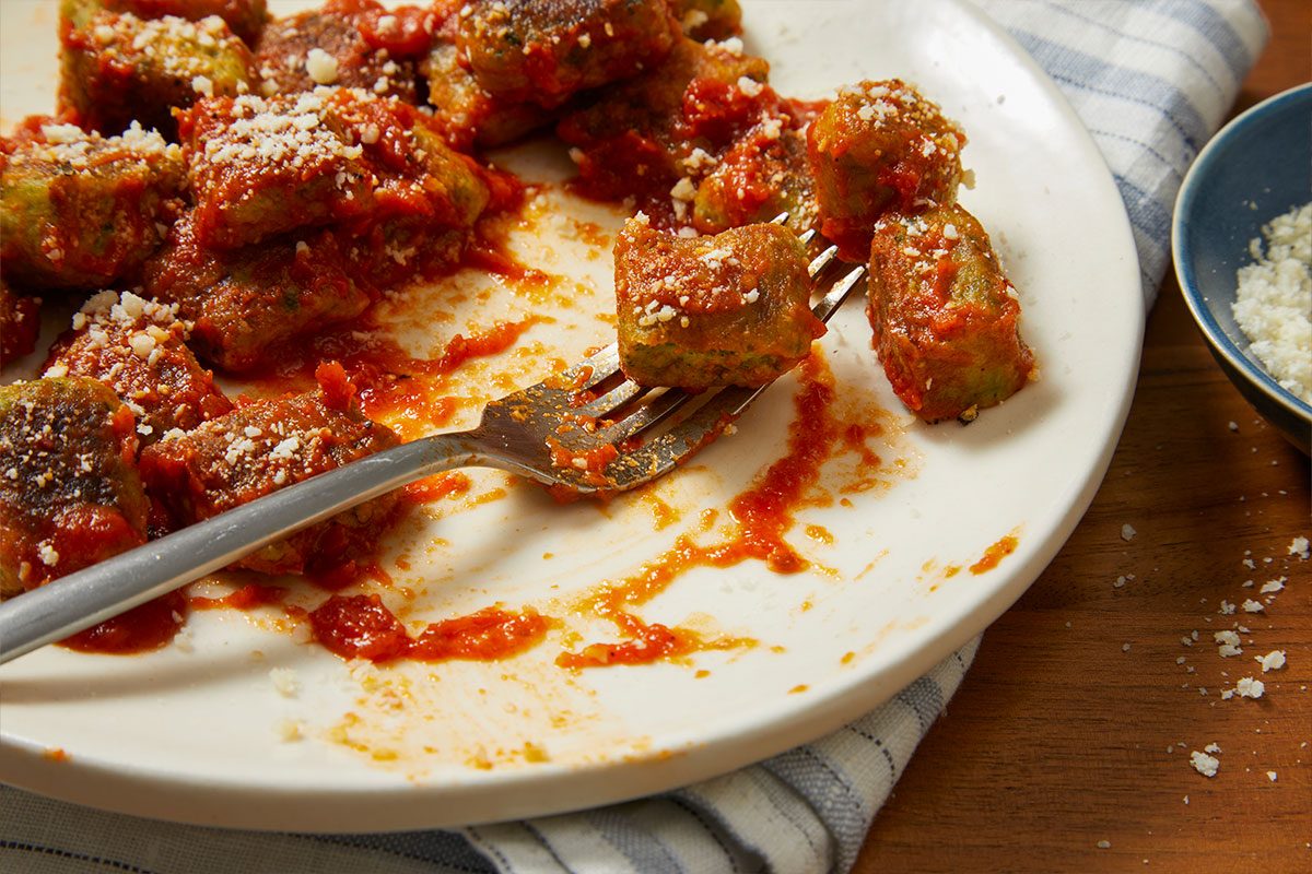 Eye Level horizontal image of gnocchi covered in red sauce and grated cheese on a white plate. A fork holds one piece of gnocchi, and a bowl of extra grated cheese is visible beside the plate.