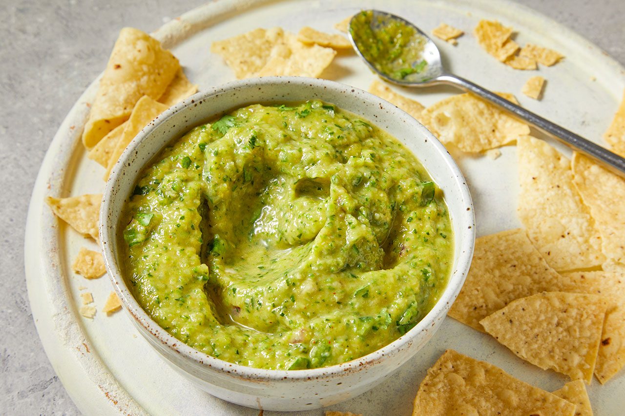 Overhead horizontal image of a bowl filled with a vibrant green dip. The chunky dip, appearing to be a type of salsa or guacamole with visible herbs like cilantro, is served alongside a scattering of tortilla chips on a light-colored tray. A spoon, also coated in the dip, rests near the bowl.