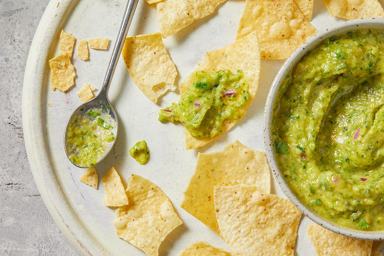 Overhead horizontal image of a bowl of chunky guacamole, surrounded by tortilla chips on a light-colored serving platter. A spoon rests beside the bowl, holding some of the green dip with visible red onion and cilantro pieces. One chip is topped with a generous serving of guacamole, ready for snacking.