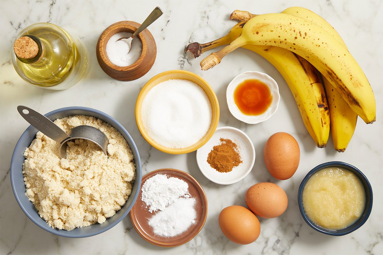 Baking ingredients on a marble surface, including bananas, eggs, flour, sugar, cinnamon, baking powder, vanilla, applesauce, oil, and salt, arranged in bowls and measuring cups.