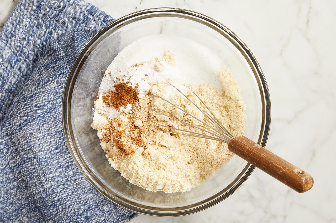 A glass mixing bowl with flour, sugar, and cinnamon being whisked together sits on a marble countertop next to a folded blue cloth.
