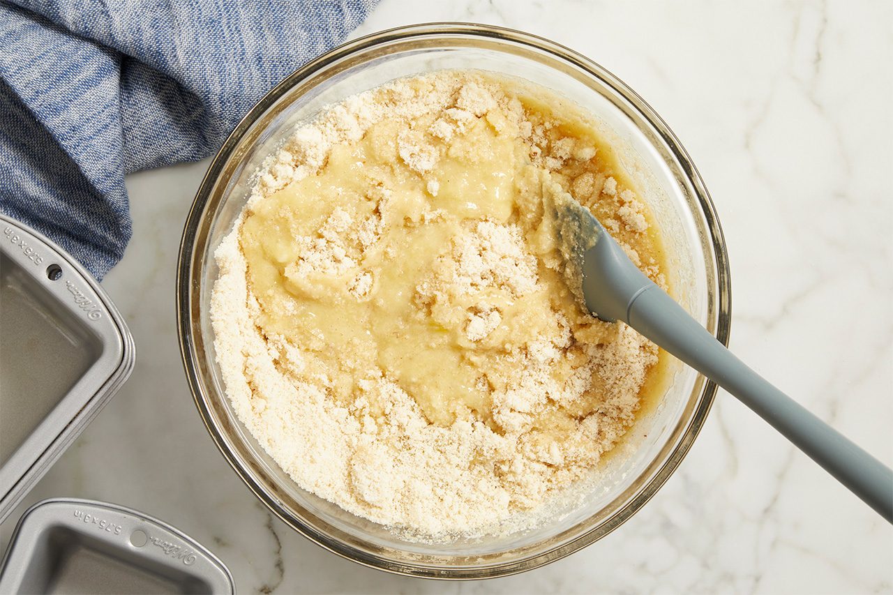 A glass bowl with flour and wet ingredients being mixed by a spatula, sitting on a marble surface next to a blue cloth and metal baking pans.