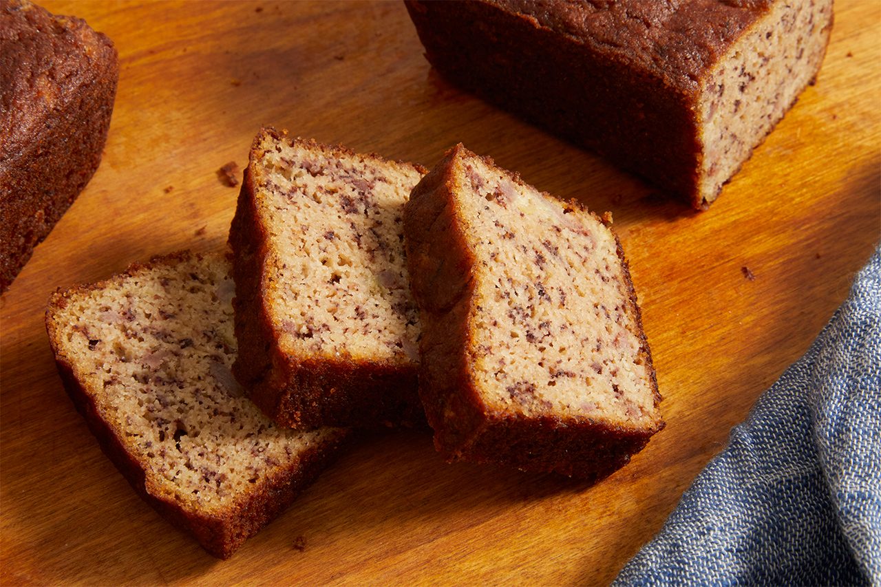 Three slices of banana bread are arranged on a wooden surface, with more banana bread loaves nearby. A portion of a blue-gray cloth is visible in the lower right corner.