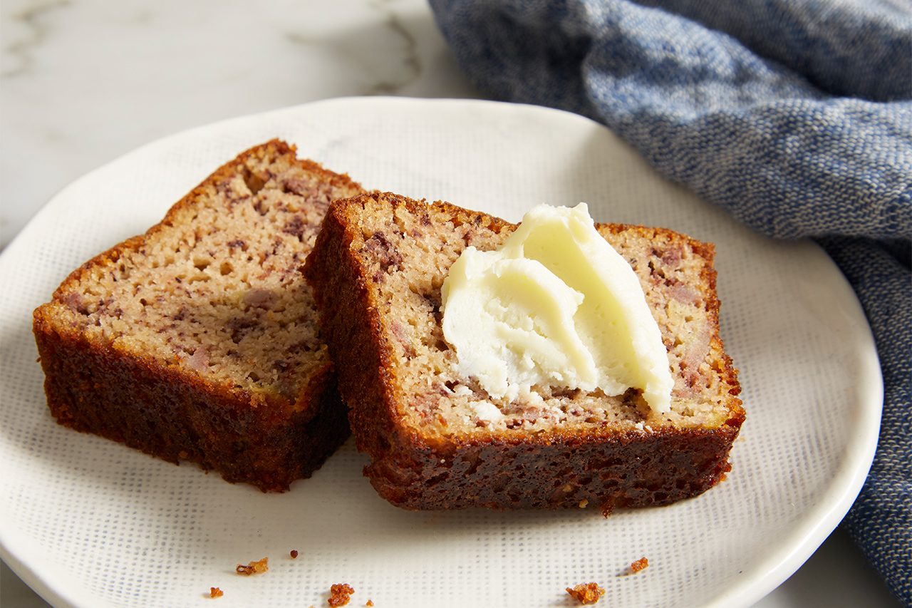 Two slices of banana bread on a white plate, with a pat of butter melting on top of one slice. A blue cloth is partially visible in the background.