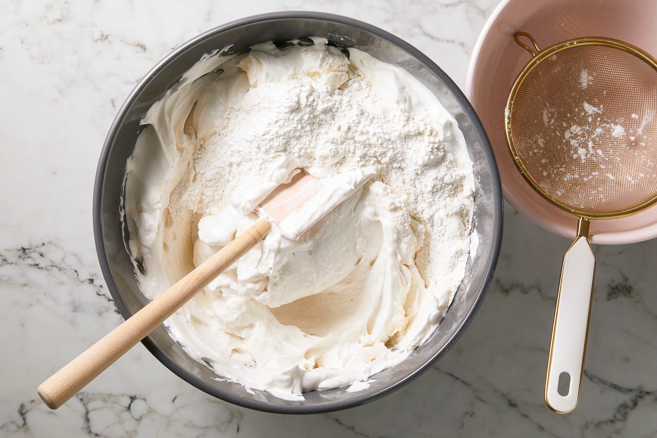 Overhead horizontal image of a metal mixing bowl filled with creamy white mixture, topped with a pile of white flour.
