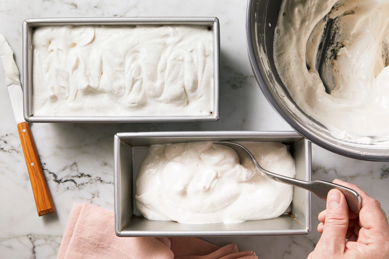 Overhead horizontal image of a person scooping a thick, white creamy mixture from a metal mixing bowl into a metal loaf pan.