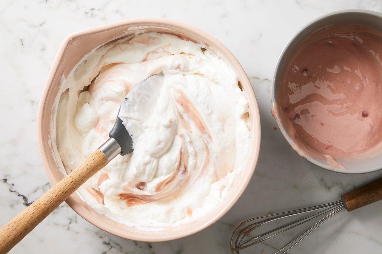 Overhead horizontal image of a pale pink bowl containing white whipped cream being swirled with a spatula, revealing streaks of a reddish-pink fruit mixture.