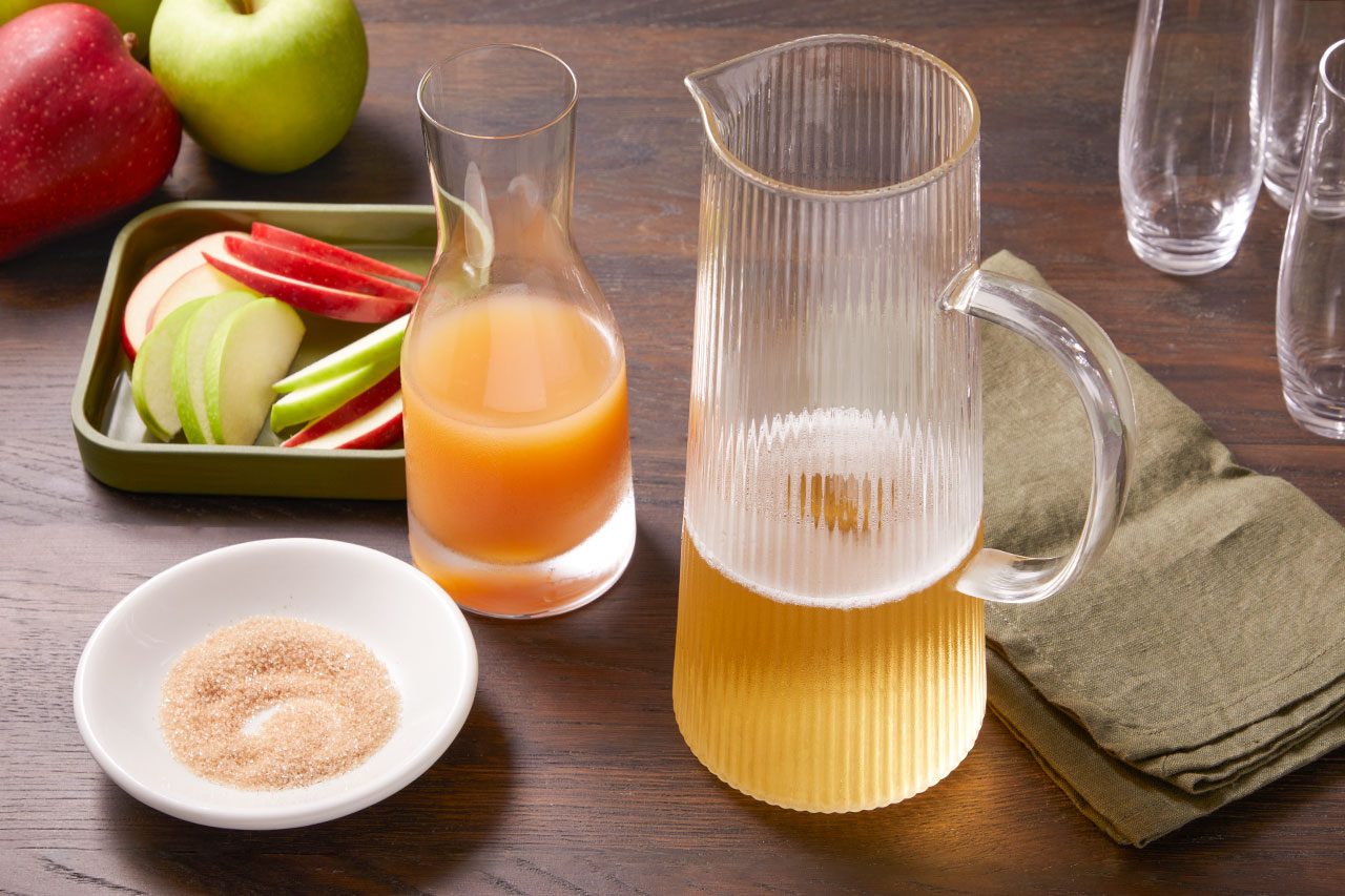 Eye level horizontal image of a tall ribbed glass pitcher with a foamy golden beverage and a smaller glass carafe filled with apple cider, set on a dark wooden surface. A small white bowl of brown sugar is visible, along with a green tray holding sliced red and green apples, whole apples, and empty glassware in the background.