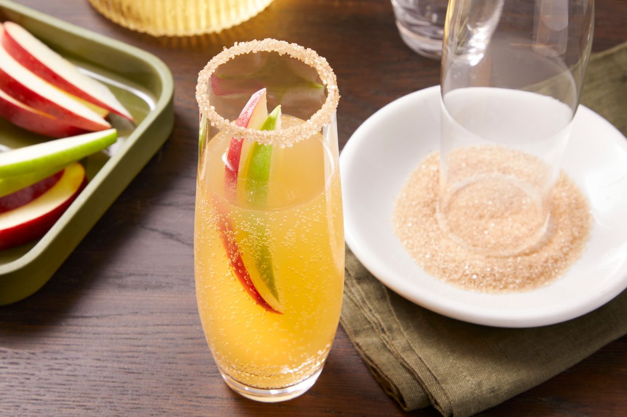Eye Level horizontal image of a sparkling drink in a tall glass, garnished with red and green apple slices and a brown sugar rim. To the right, a white bowl holds brown sugar with an empty glass inside, while a green tray to the left contains additional red and green apple slices.