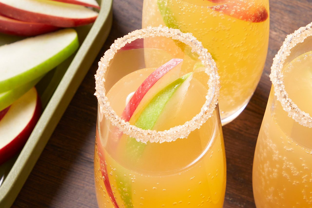 Eye Level horizontal image of multiple glasses filled with a bubbly, golden beverage. The glasses have sugared rims and are garnished with red and green apple slices, with more apple slices visible on a tray in the background.