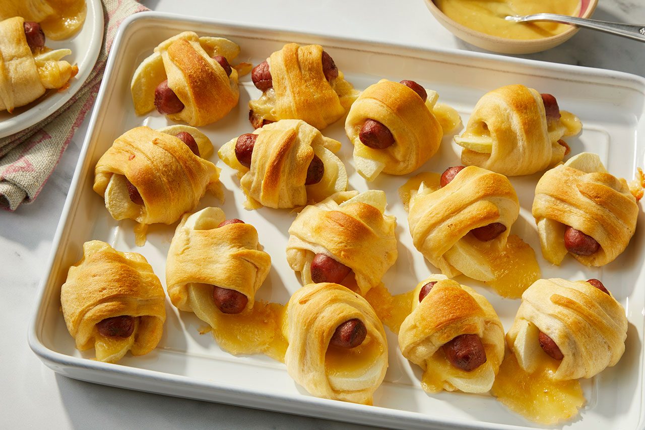 Overhead horizontal image of numerous freshly baked Apple-Gouda Pigs in a Blanket arranged in a white rectangular baking dish. The golden brown crescent rolls encase miniature smoked sausages, apple slices, and melted Gouda cheese, with some cheese oozing out. A small bowl of honey mustard salad dressing is visible in the background.