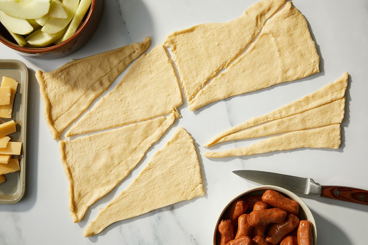 Overhead horizontal image of crescent roll dough triangles laid out on a white marble surface, next to bowls of sliced apples, miniature smoked sausages, and cubed Gouda cheese. A paring knife is also visible near the sausages, indicating preparation for Apple-Gouda Pigs in a Blanket.