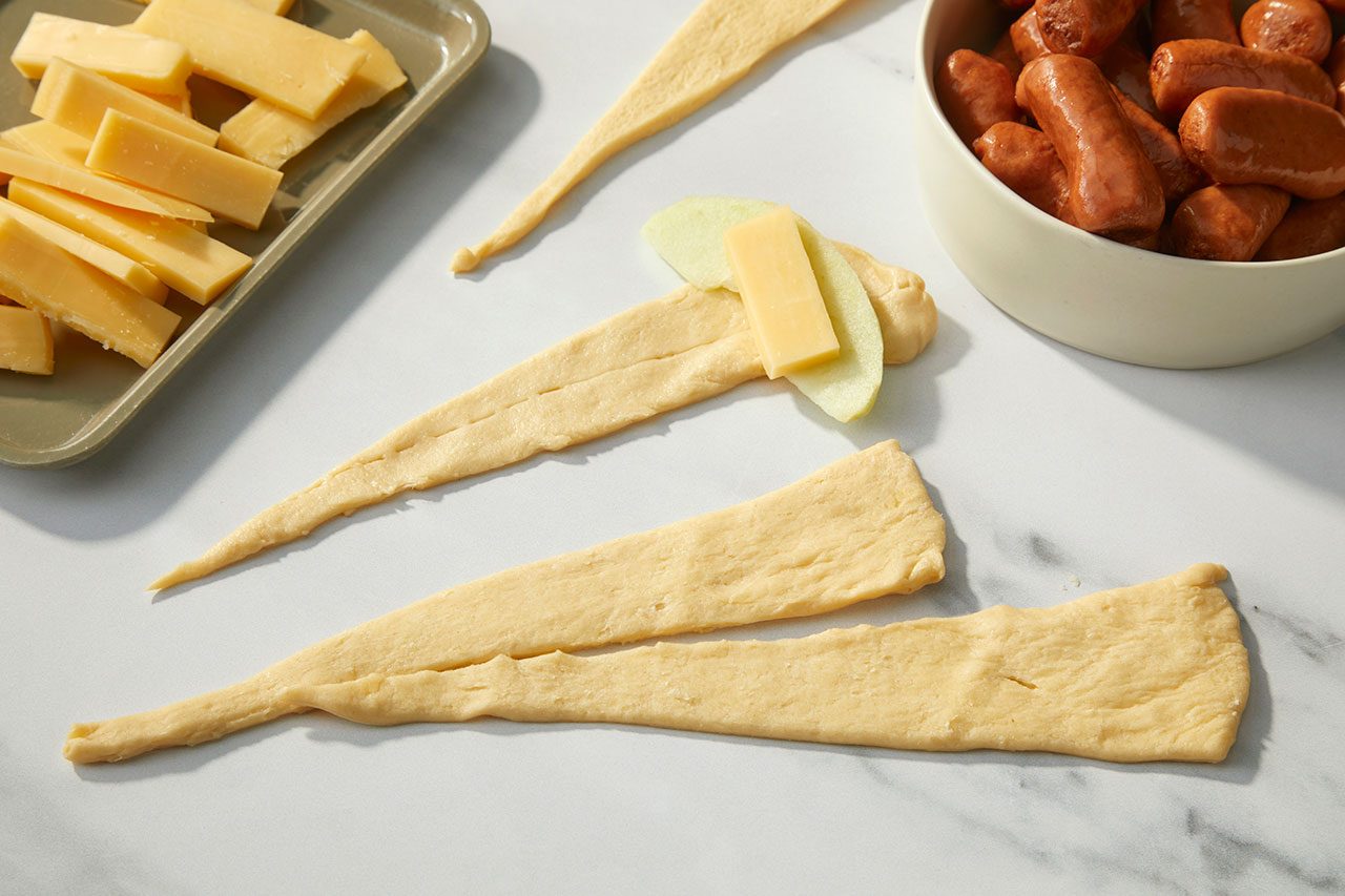 Overhead horizontal image of several crescent roll dough triangles on a white marble surface. One dough triangle has thin apple slices and a piece of Gouda cheese placed on it, ready to be rolled. A gray tray of Gouda cheese sticks and a white bowl of miniature smoked sausages are visible in the background.