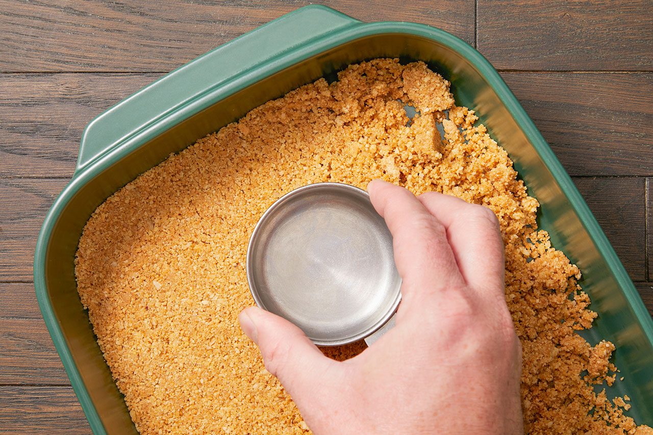 Overhead horizontal image of a hand pressing down a light brown crumb mixture into the bottom of a dark green rectangular baking dish. The hand uses a small, round metal cup to compact the crumbs evenly across the pan, forming a crust.