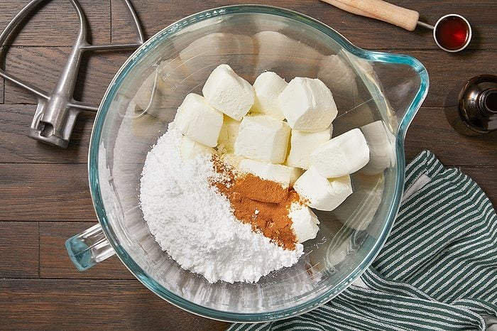 Overhead horizontal image of ingredients being combined in a clear glass mixing bowl. Cubes of cream cheese, powdered sugar, and cinnamon are visible in the bowl, ready to be mixed. A stand mixer paddle and vanilla extract are visible nearby.