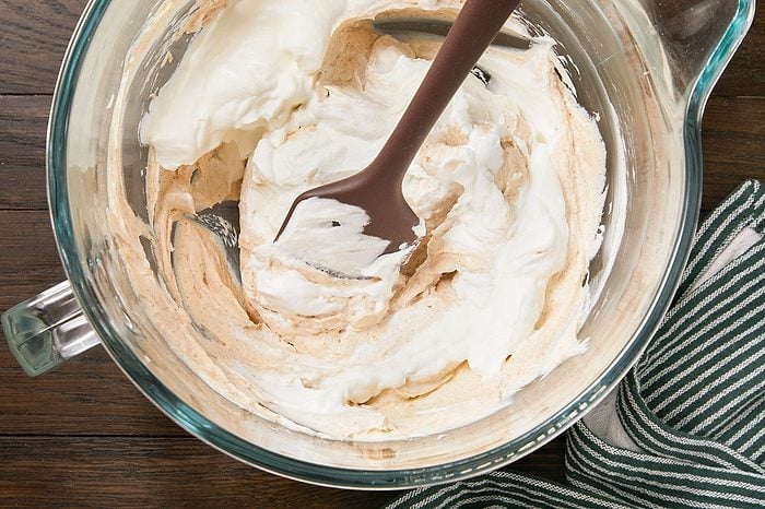 Overhead horizontal image of a brown spatula mixing a creamy, light brown mixture with a white, fluffy ingredient in a clear glass bowl. The ingredients are being folded together, creating swirls of both textures.