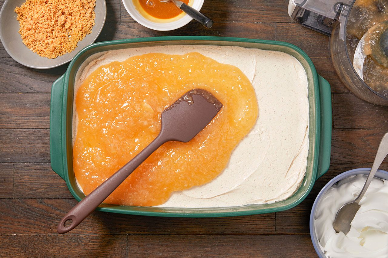 Overhead horizontal image of a brown spatula spreading a thick layer of apple compote over a creamy, light-colored dessert base in a green baking dish. Surrounding ingredients include a bowl of crushed crumbs, a bowl of caramel sauce, and a bowl of whipped cream.