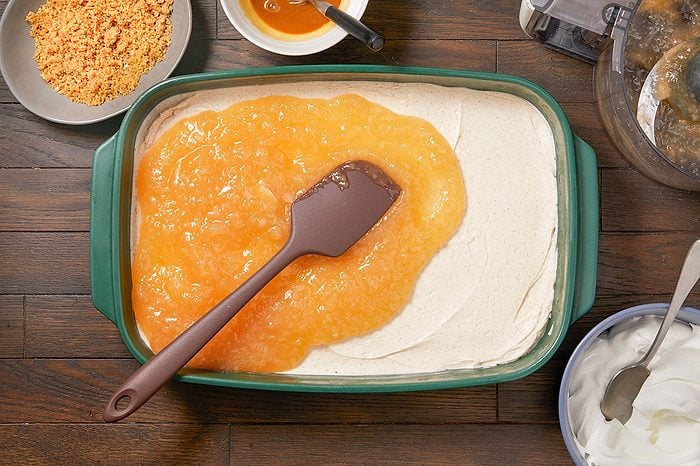 Overhead horizontal image of a brown spatula spreading a thick layer of apple compote over a creamy, light-colored dessert base in a green baking dish. Surrounding ingredients include a bowl of crushed crumbs, a bowl of caramel sauce, and a bowl of whipped cream.