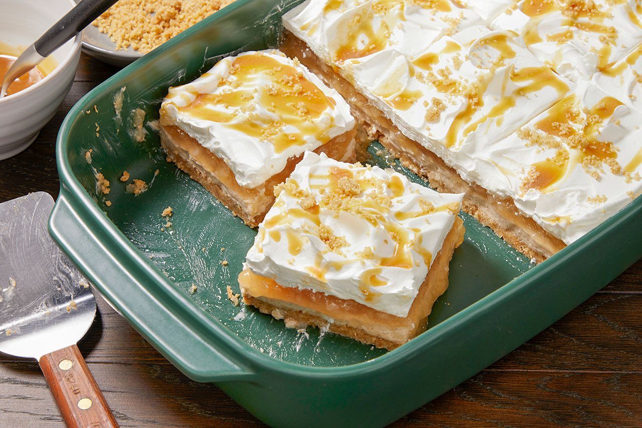 Eye Level horizontal image of a green baking dish holding several slices of a layered dessert. The dessert features a bottom crust, a creamy filling, and a generous topping of whipped cream, drizzled with caramel sauce and sprinkled with crushed crumbs. A serving spatula is visible next to the dish, with additional caramel sauce and crumbs in the background.