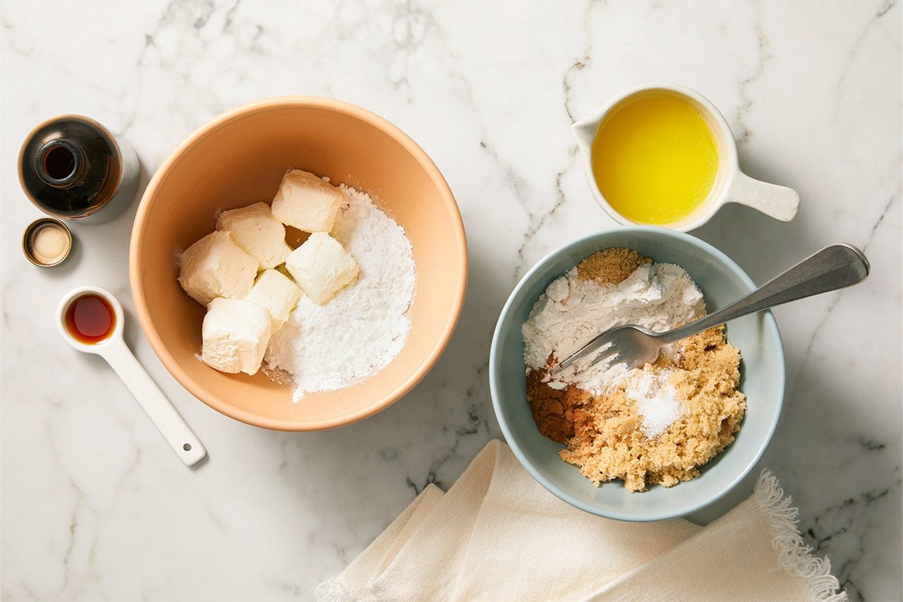 Overhead horizontal image of baking ingredients laid out on a white marble surface. Cubed cream cheese and powdered sugar are combined in a tan bowl, while a blue bowl contains flour, brown sugar, cinnamon, and salt with a fork. Melted butter in a measuring cup and vanilla extract are also visible.