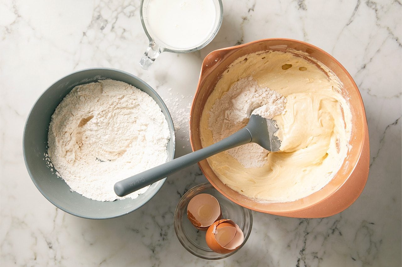 Overhead horizontal image of ingredients being prepared for a recipe. A grey bowl holds white flour, while a brown mixing bowl contains yellow batter with additional flour and a grey spatula. A glass measuring cup with milk and a small bowl with empty eggshells are also visible on a marble surface.