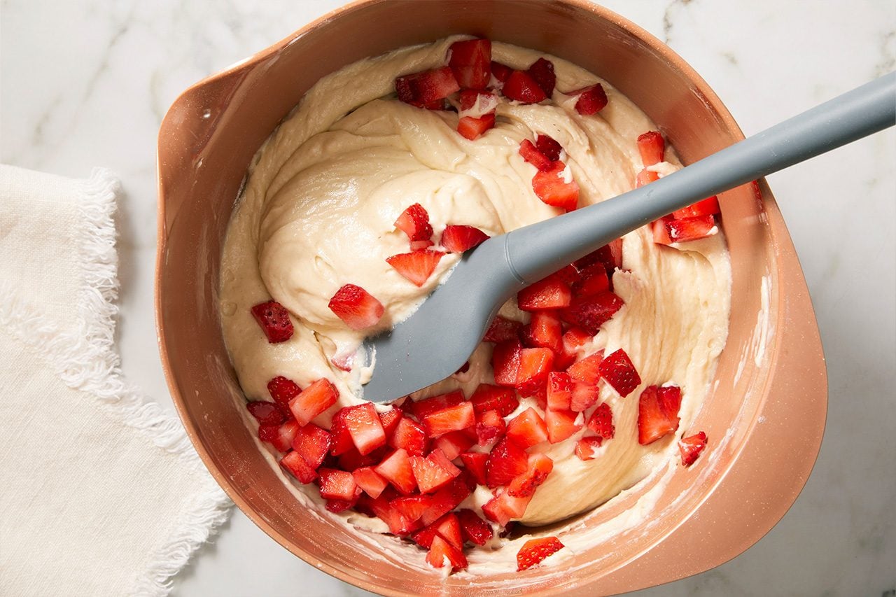 Overhead horizontal image of diced strawberries being folded into a light-colored batter within a rose gold-colored mixing bowl. A grey spatula is partially submerged, aiding in the mixing process. The bowl sits on a white marble surface next to a white linen napkin.