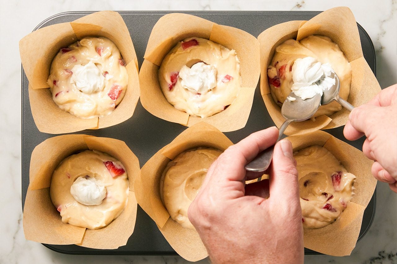 Overhead horizontal image of hands spooning a dollop of white cream cheese filling into strawberry muffin batter. The batter, with visible red strawberry pieces, rests in brown paper liners within a gray muffin tin.