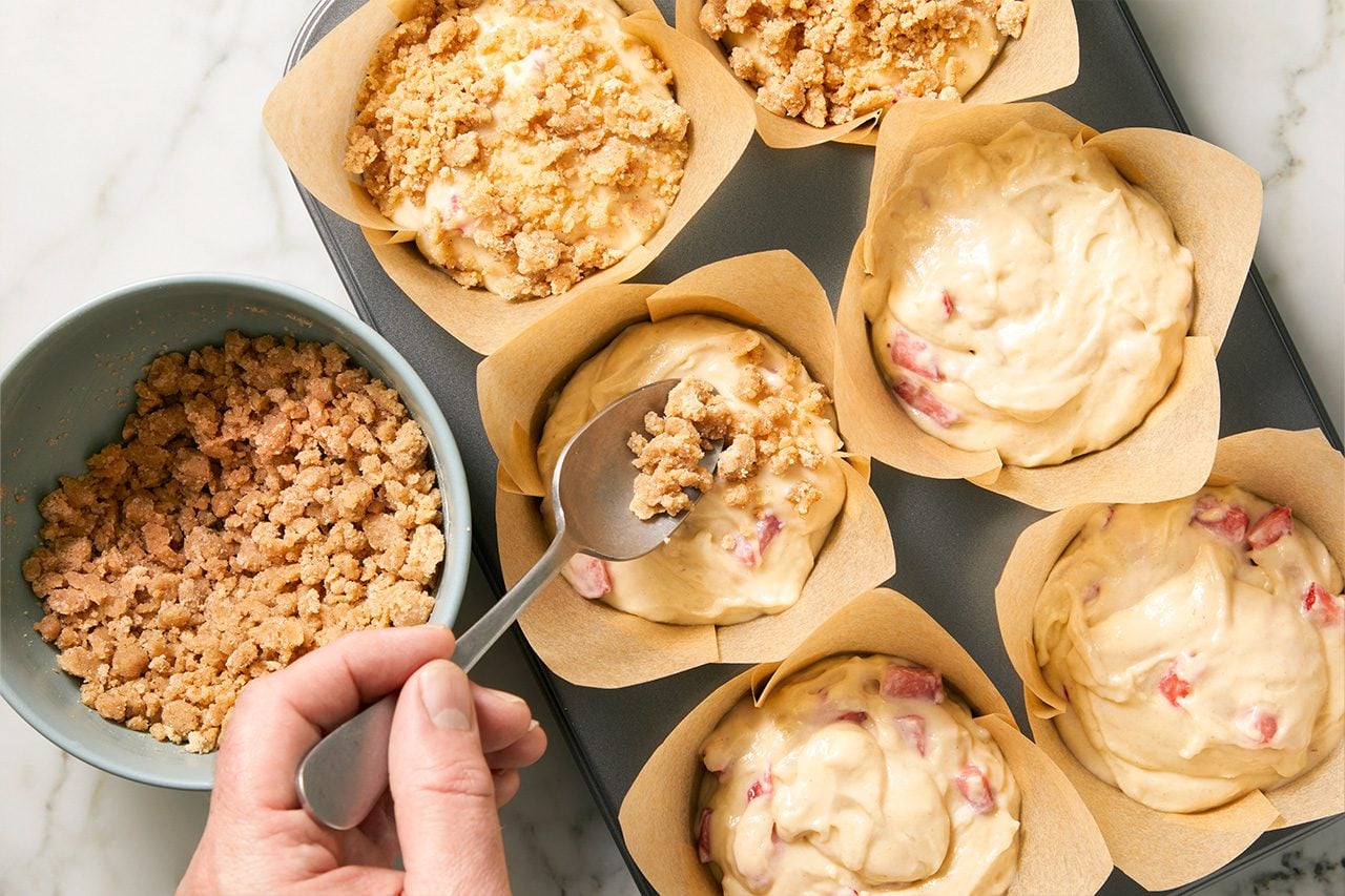 Overhead horizontal image of a hand using a spoon to add a generous crumb topping to muffin batter. The batter, which contains visible red fruit pieces, is spooned into paper liners within a muffin tin. A bowl filled with more crumb topping is visible to the left, ready for application.