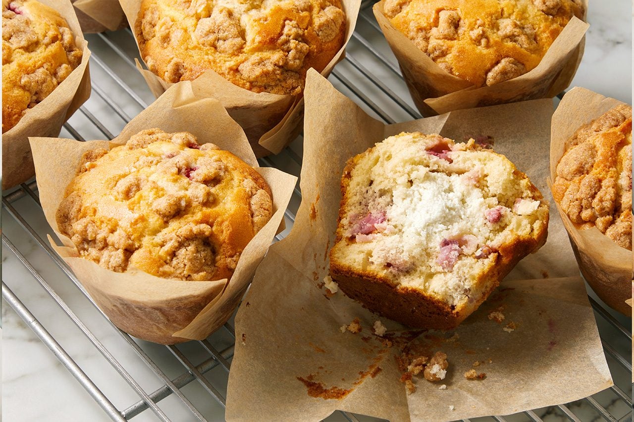 Eye level horizontal image of several freshly baked muffins cooling on a wire rack. One muffin, still in its paper liner, is cut in half, revealing a fluffy interior studded with fruit pieces and a creamy white filling. All muffins are topped with a golden-brown streusel.